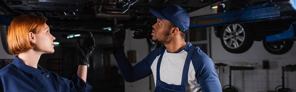 Side view of multiethnic mechanics pointing at car bottom in service, banner