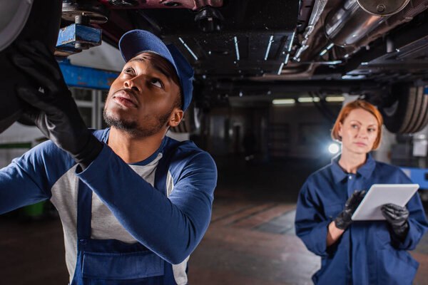 African american mechanic looking at car wheel near blurred colleague with digital tablet in service 