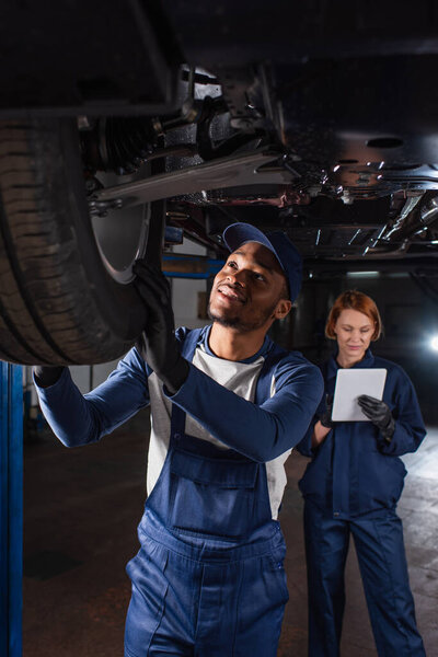 Smiling african american mechanic looking at car wheel near colleague using digital tablet in garage 