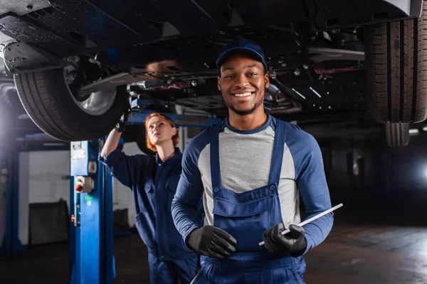Smiling african american mechanic with digital tablet looking at camera near colleague and car in garage
