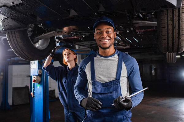 Smiling african american mechanic with digital tablet looking at camera near colleague and car in garage 