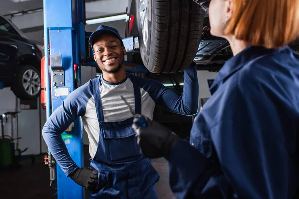 Smiling african american mechanic looking at camera near car and colleague in service