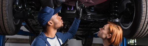 Side view of african american mechanic pointing at car bottom near colleague in garage, banner