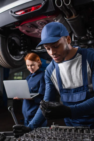 African american mechanic holding tools while colleague using laptop near car in garage