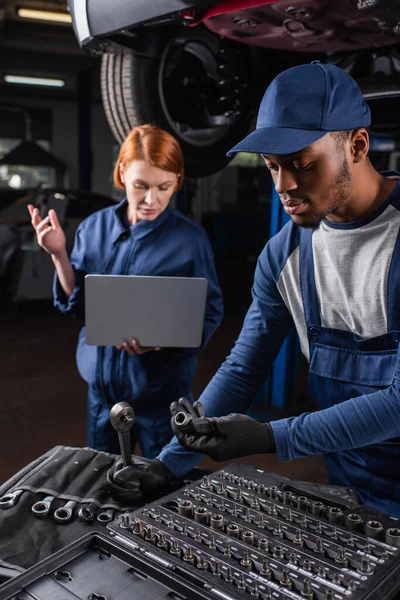 Africano Americano Mecánico Herramientas Retención Cerca Borrosa Colega Utilizando Portátil — Foto de Stock