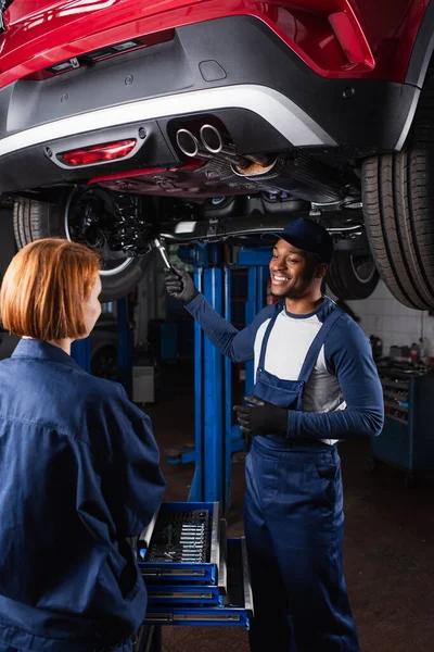 Cheerful african american mechanic holding wrench near car bottom and colleague in service