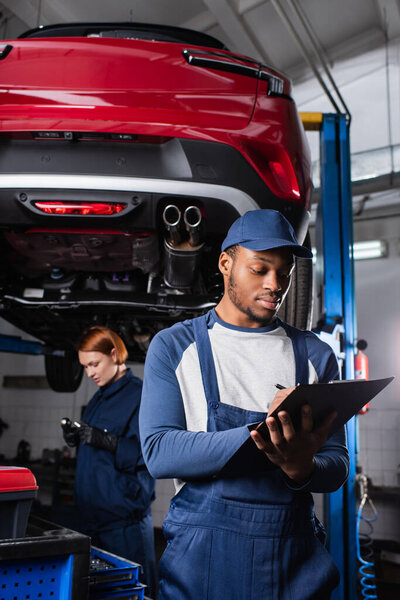 African american mechanic in uniform writing on clipboard near colleague and auto in garage 