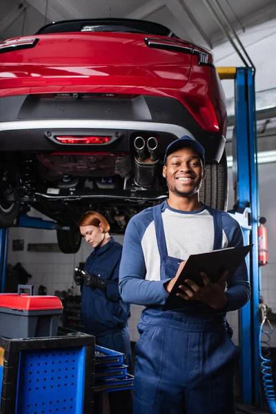 Smiling african american mechanic writing on clipboard and looking at camera in car service