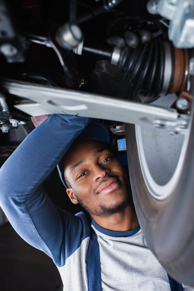 african american repairman smiling at camera while inspecting shock absorber of car