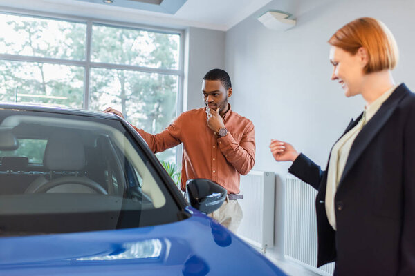 thoughtful african american man looking at car near blurred car dealer