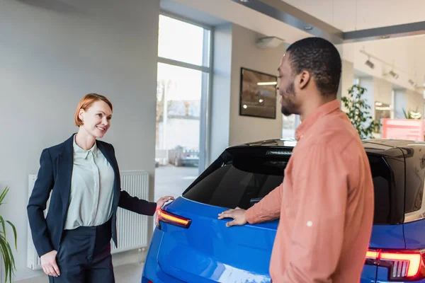 快乐的汽车经销商和模糊的非洲裔美国客户站在展示厅的汽车旁边 — 图库照片