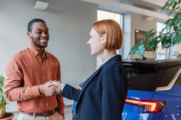 happy african american man and car dealer shaking hands near car 