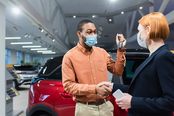 車のショールームで握手する医療マスクのキーと車のディーラーを持つアフリカ系アメリカ人男性 — ストック写真