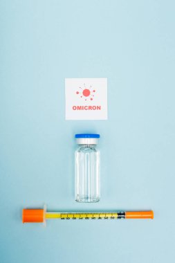 top view of white card with omicron lettering and bacteria near vaccine vial and syringe on blue