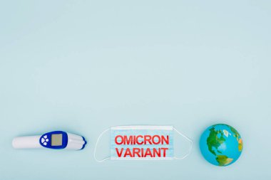top view of medical mask with red omicron variant lettering near globe and pyrometer on blue