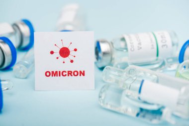white card with red omicron lettering and bacteria near blurred vaccine vials and ampoules on blue
