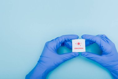 white card with red omicron lettering and bacteria icon in hands of doctor in latex gloves on blue, cropped view