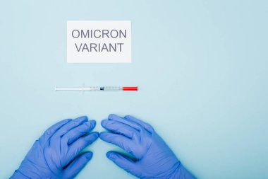 top view of doctor in latex gloves near card with omicron variant lettering and syringe on blue