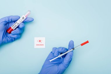 top view of doctor in latex gloves holding syringe and test tube near card with omicron lettering and bacteria on blue