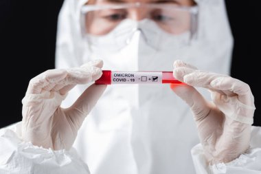 blurred doctor in hazmat suit holding positive covid-19 omicron variant test isolated on black