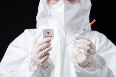 partial view of doctor in hazmat suit holding syringe and card with omicron lettering and bacteria icon isolated on black
