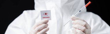 partial view of scientist in latex gloves holding syringe and card with omicron lettering isolated on black, banner