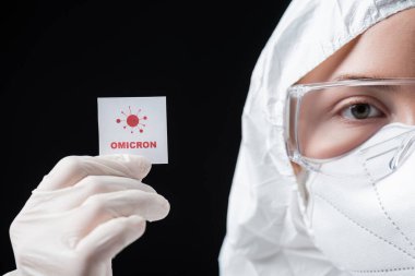 cropped view of doctor in hazmat suit holding card with omicron lettering and bacteria icon isolated on black