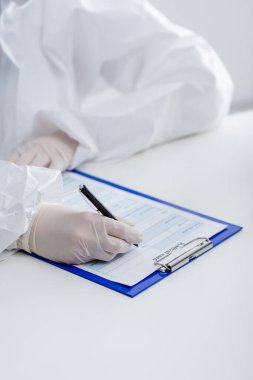 partial view of blurred doctor in hazmat suit writing in medical card on clipboard in lab