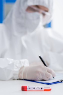 blurred scientist in hazmat suit writing on clipboard near positive covid-19 omicron variant test in lab