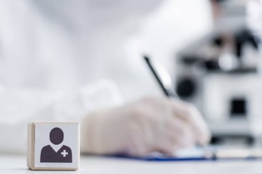 selective focus of cube with medical icon near scientist writing near microscope on blurred background, omicron variant concept