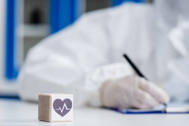 blurred scientist in personal protective equipment writing on clipboard near cube with heart sign