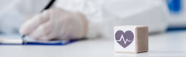 partial view of blurred doctor writing on clipboard near cube with heart sign, banner