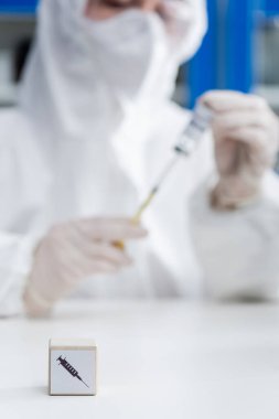 cropped view of blurred immunologist in hazmat suit holding vaccine near cube with syringe icon, omicron variant concept