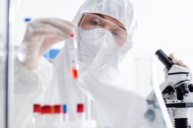 scientist in goggles, medical mask and hazmat suit holding blurred test tube near microscope in lab