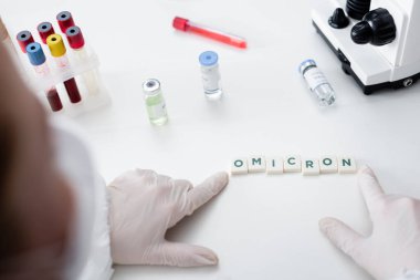 partial view of blurred scientist near cubes with omicron lettering, vaccine vials and test tubes in lab
