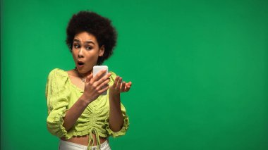 Şok olmuş Afro-Amerikalı kadın yeşil alanda izole edilmiş akıllı telefon kullanıyor. 