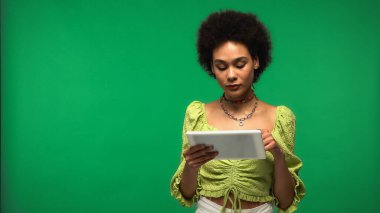 Bluzlu kıvırcık Afrikalı Amerikalı kadın yeşil üzerine izole edilmiş dijital tablet kullanıyor. 