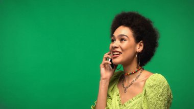 Bluz giymiş neşeli Afro-Amerikan kadın yeşil alanda izole edilmiş akıllı telefondan konuşuyor. 