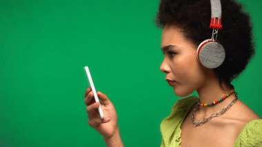 side view of african american woman in wireless headphones listening music and using smartphone isolated on green 