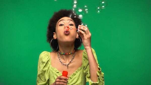 curly african american woman blowing soap bubbles while holding bubble wand isolated on green 