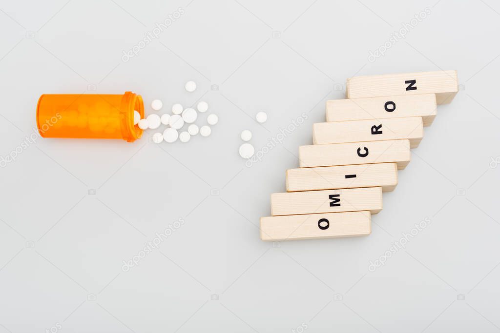 top view of container and pills near wooden bricks with omicron lettering on grey background