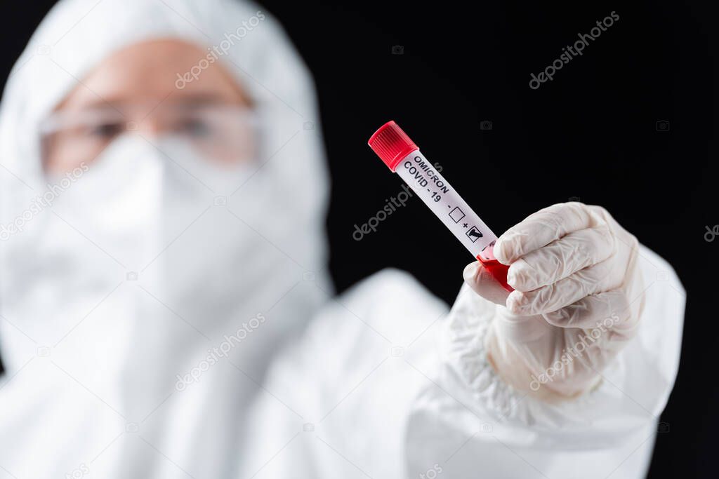 blurred scientist in white hazmat suit holding positive covid-19 omicron variant test isolated on black