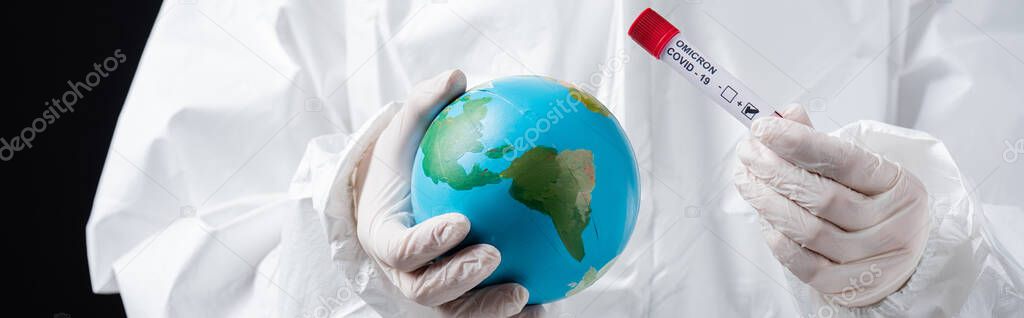 cropped view of doctor in hazmat suit holding globe and covid-19 test tube isolated on black, banner