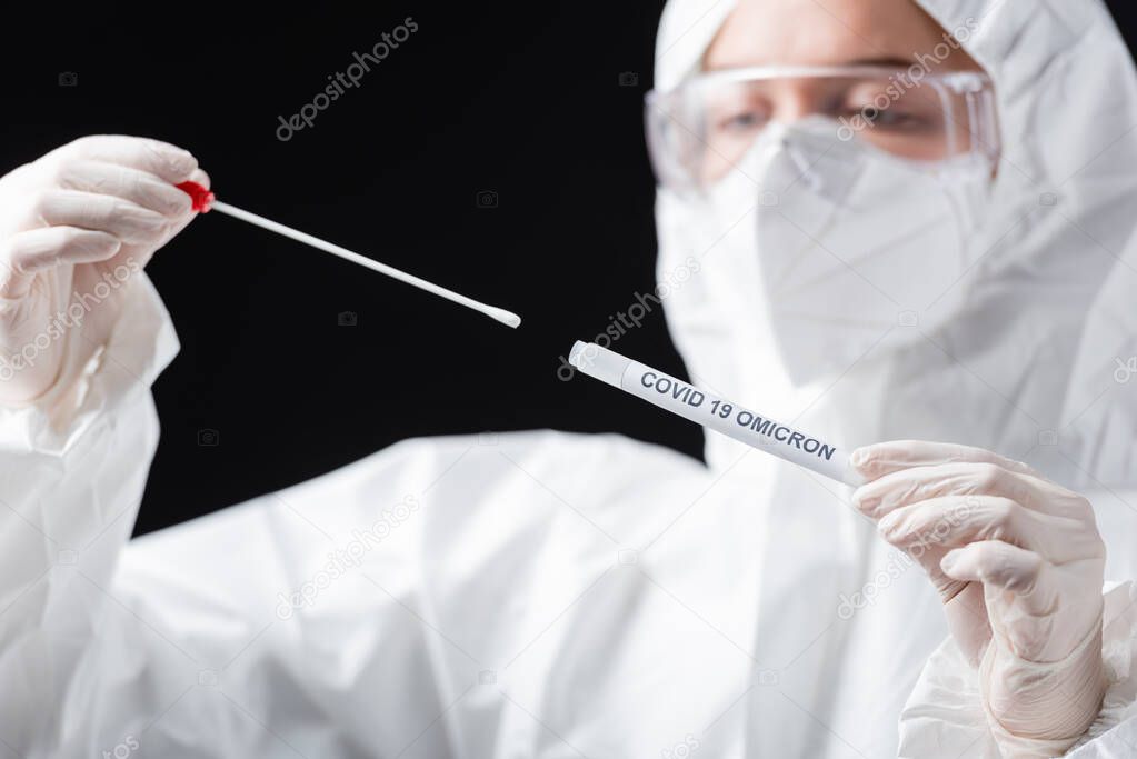 blurred scientist in hazmat suit holding covid-19 omicron variant swab test isolated on black