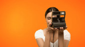 Young woman taking photo on vintage camera isolated on orange 