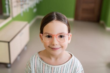 happy girl in eyeglasses looking at camera in optics store on blurred background