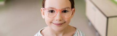 cheerful girl in eyeglasses smiling at camera in optics store, banner