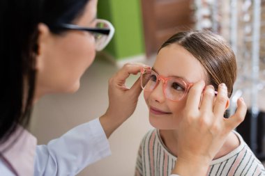 blurred oculist trying eyeglasses on smiling girl in optics shop