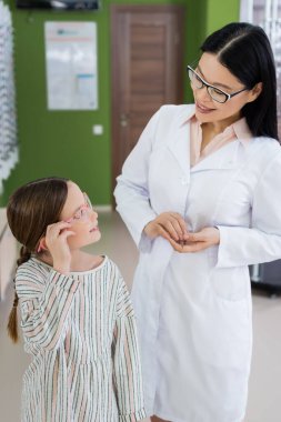asian oculist smiling at girl trying on eyeglasses in optics shop