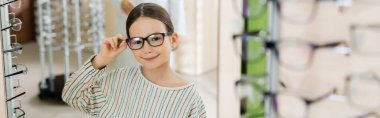 happy girl looking at camera while choosing eyeglasses in optics shop on blurred foreground, banner